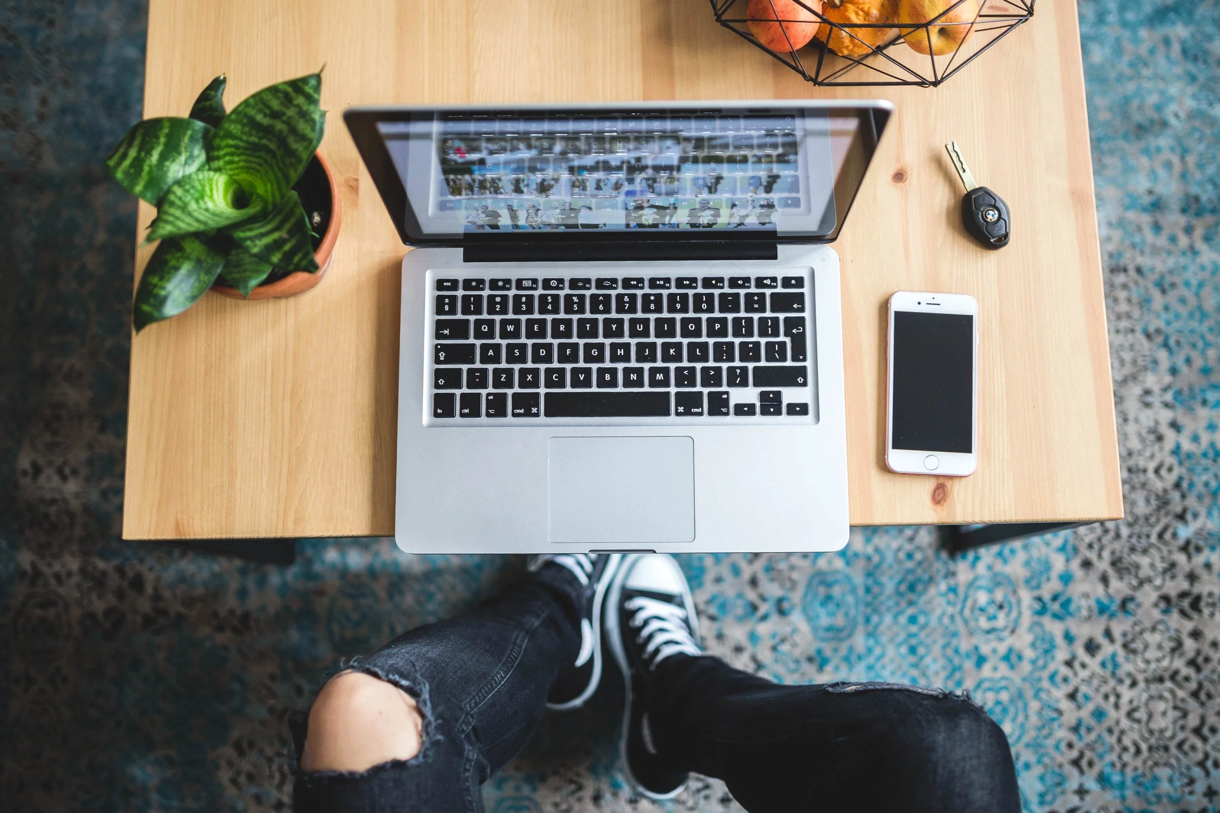 Top-down view of a workspace with a laptop, smartphone, car key, and a plant on a wooden desk, with a person wearing black ripped jeans and black sneakers standing below.