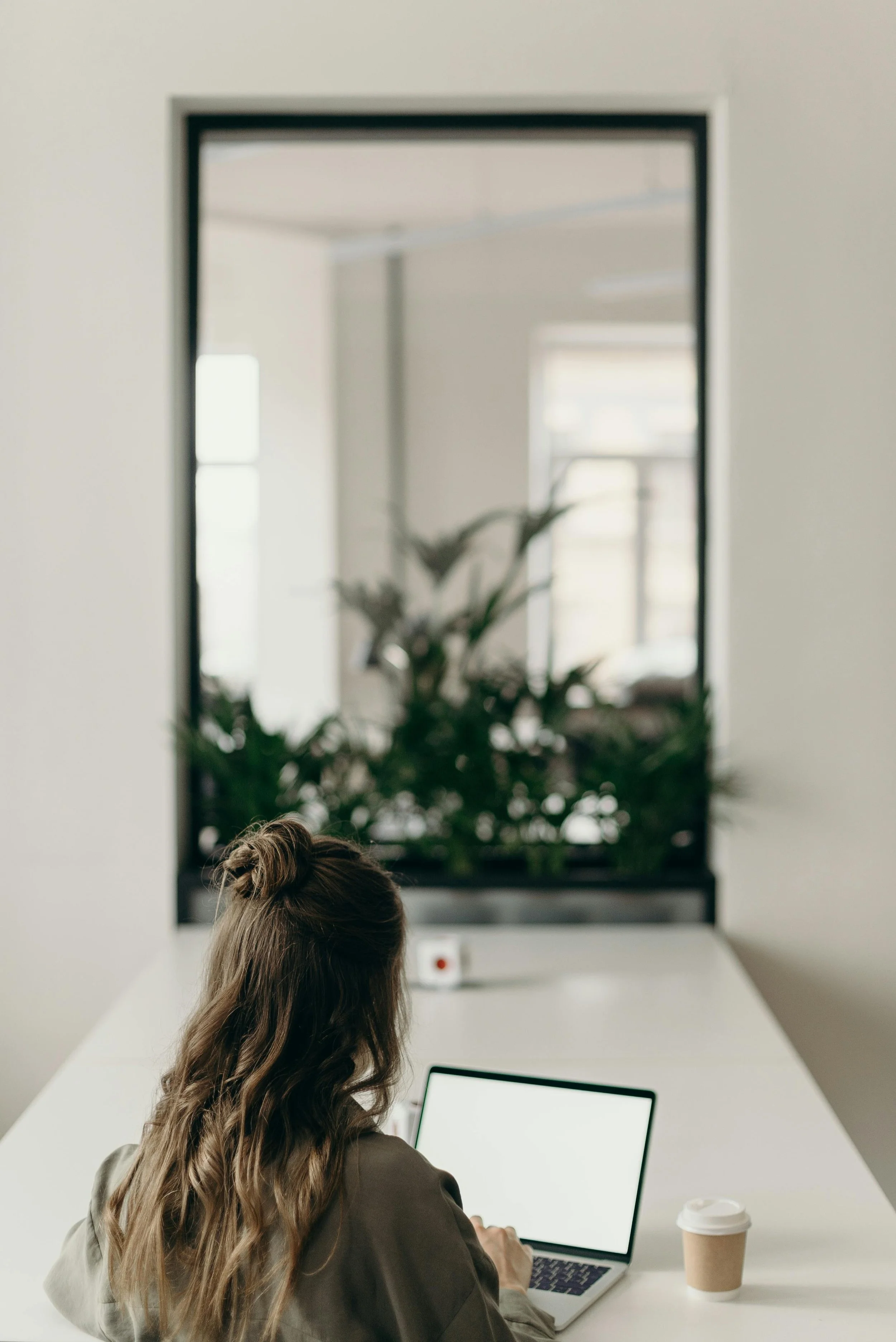 A woman with wavy hair tied back sitting at a white table working on a laptop, with a paper coffee cup beside her, in a modern office with a large window and a mirror reflecting an indoor plant.
