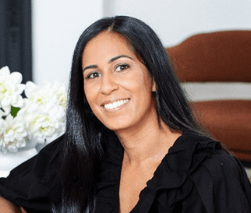 A smiling woman with long black hair wearing a black top, sitting indoors with a white flower arrangement and a brown headboard in the background.
