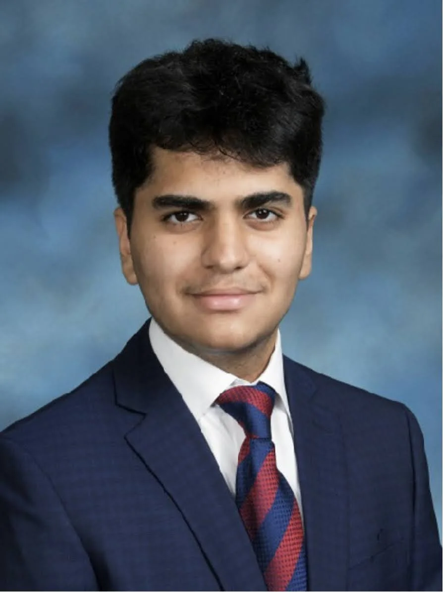 A young woman with shoulder-length black hair smiling and showing her teeth, standing among green plants.