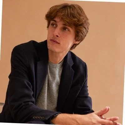 A man with glasses and a nose ring sitting indoors, wearing a dark green sweater, with a blurred background and a plant to the right.