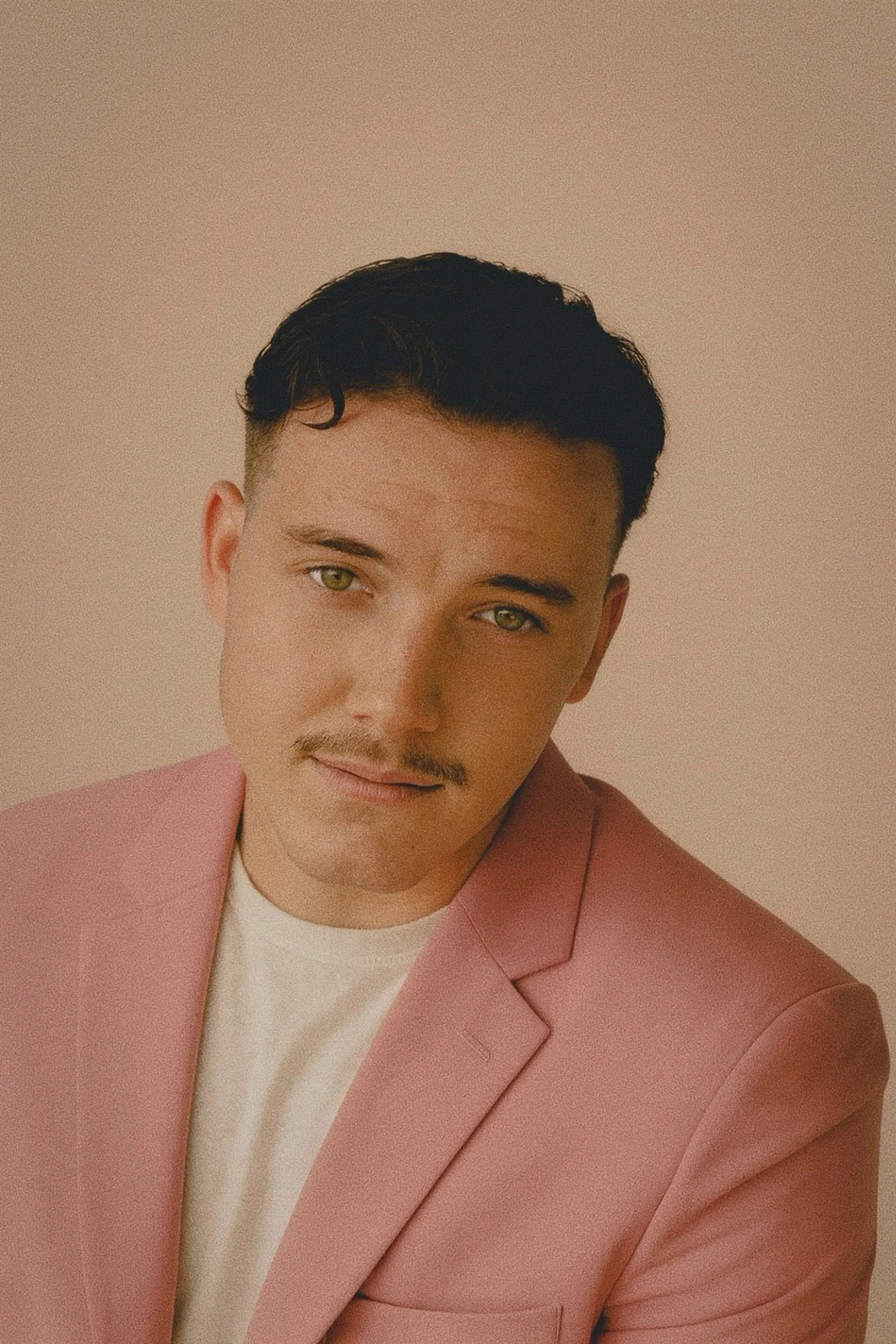 A young man with dark hair, light skin, and a mustache, wearing a pink blazer and white shirt, posing against a neutral background.