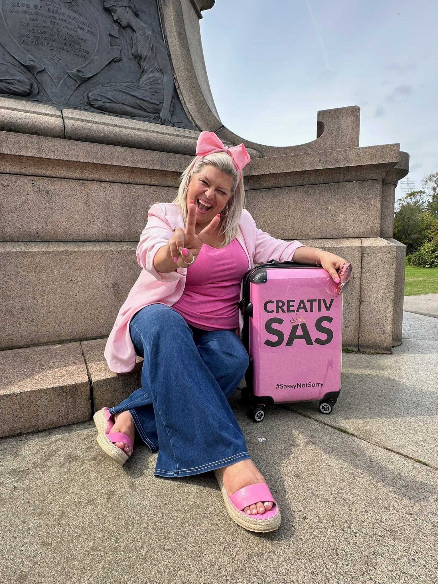 A woman sitting on a stone step next to a pink suitcase with the text 'CREATIV SAS' and '#SassyNotSorry'. She is smiling, making a peace sign, and dressed in pink and blue, with a pink bow in her hair. The background features a monument with a bronze relief sculpture.
