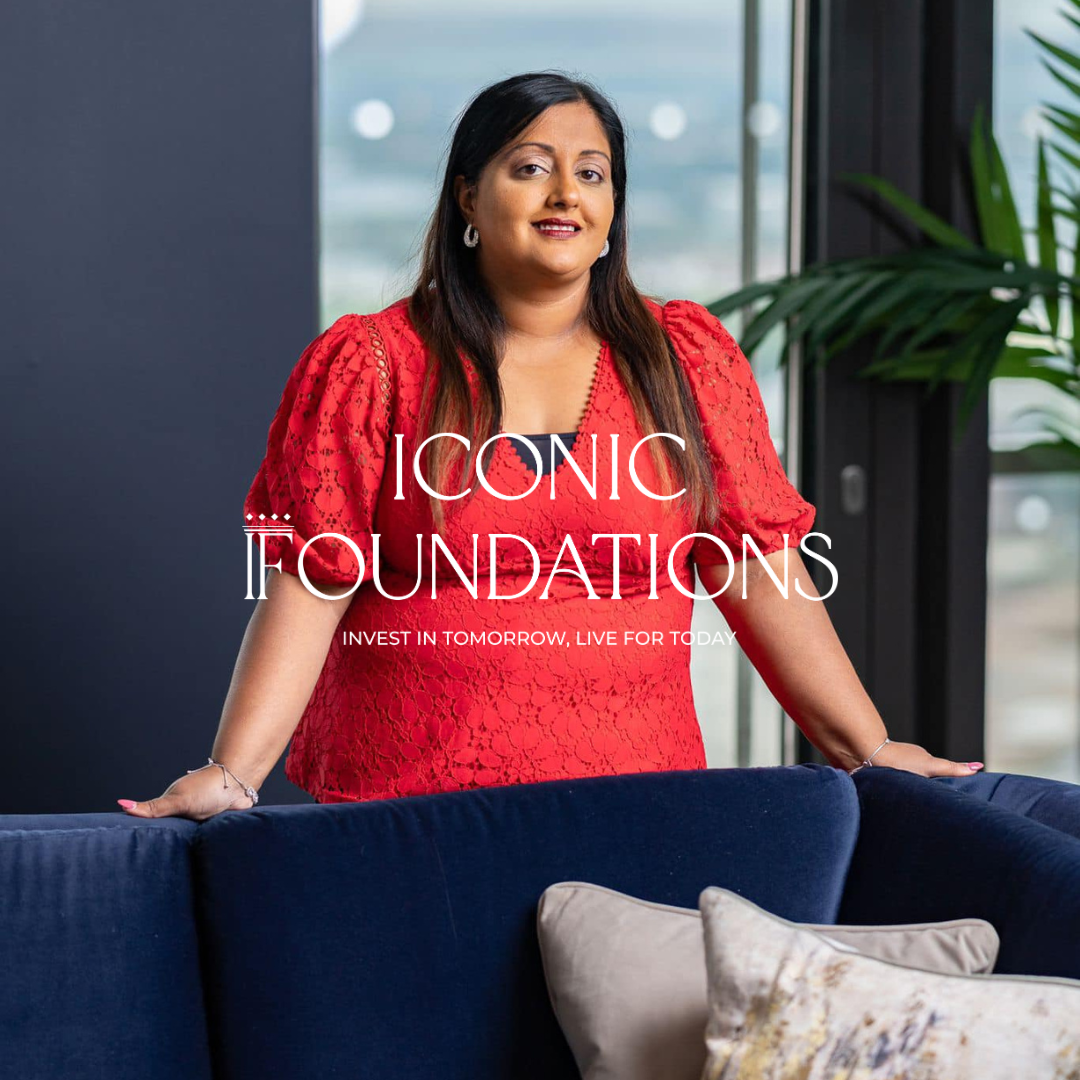 A woman in a red lace dress standing behind a navy blue sofa, smiling in a modern office with large windows and green plants in the background.