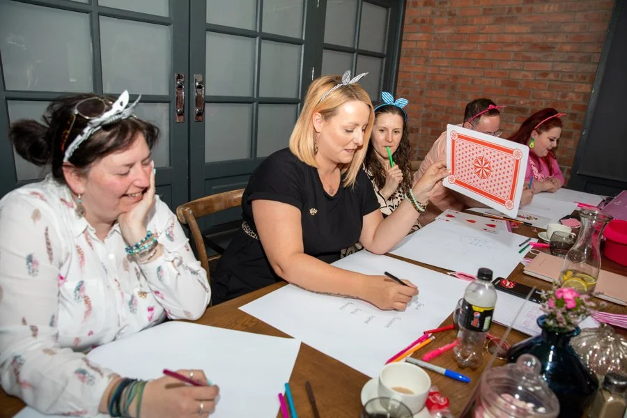 Women sitting at a table, engaging in a fun activity with one woman holding a red and white illustrated card, others wearing colorful headbands with bows, papers, pens, and drinks on the table.