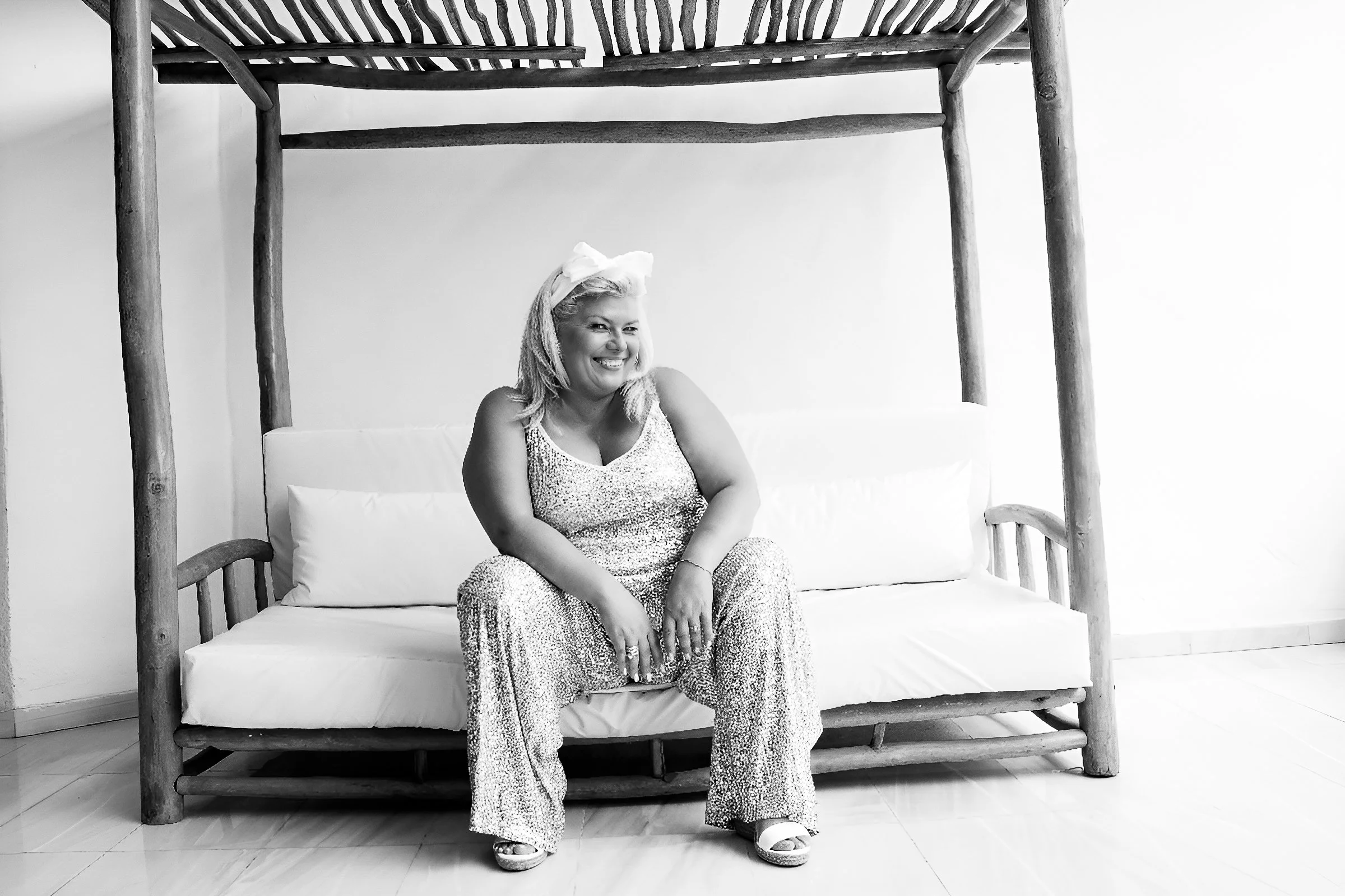 A woman sitting on a wooden bench with a cushioned seat, smiling and looking to her left. She is wearing patterned pants, a sleeveless top, and sandals. She has a bow in her hair and is in a room with a white wall and a tiled floor.