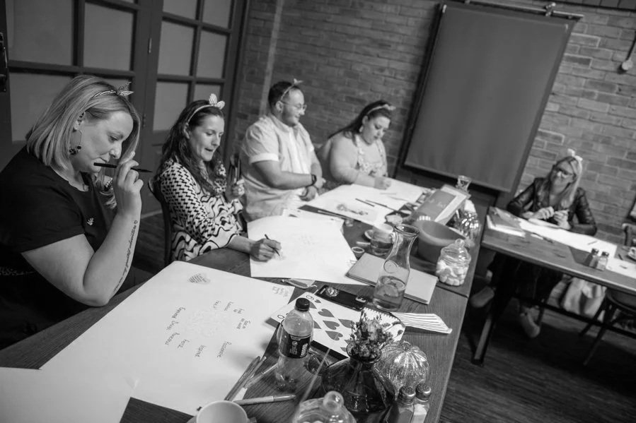 People sitting at a long table working on papers and writing, with snacks and drinks on the table, in a room with brick walls.
