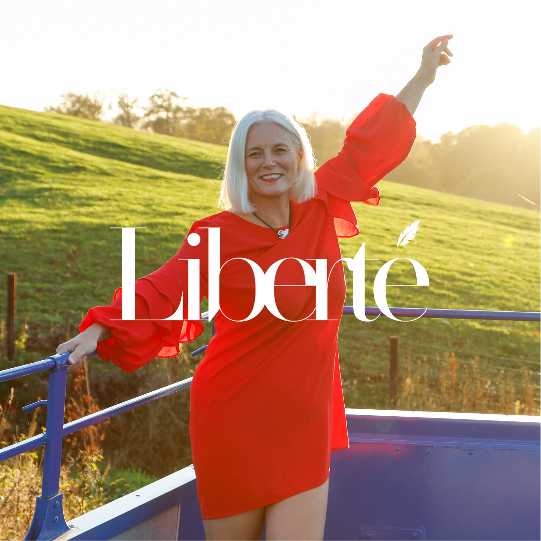 A smiling woman with white hair in a red dress standing outdoors on a bridge, with a green hill and trees in the background during golden hour sunlight.