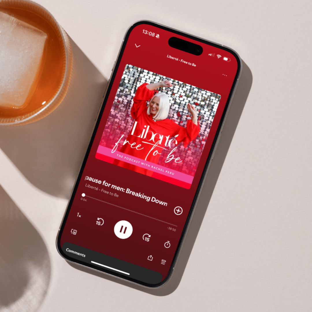Smartphone playing a podcast called "Pause for Men: Breaking Down" on a red background, with a woman smiling on the podcast cover, and a cup of coffee nearby on a white table.