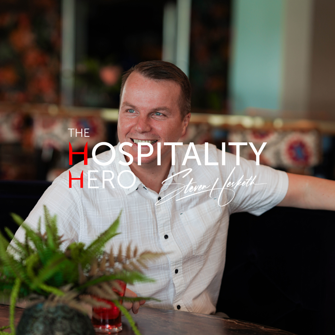 A smiling man in a white shirt sitting at a table in a restaurant with green plants, with text overlay that reads 'The Hospitality Hero Steven Hesketh'.