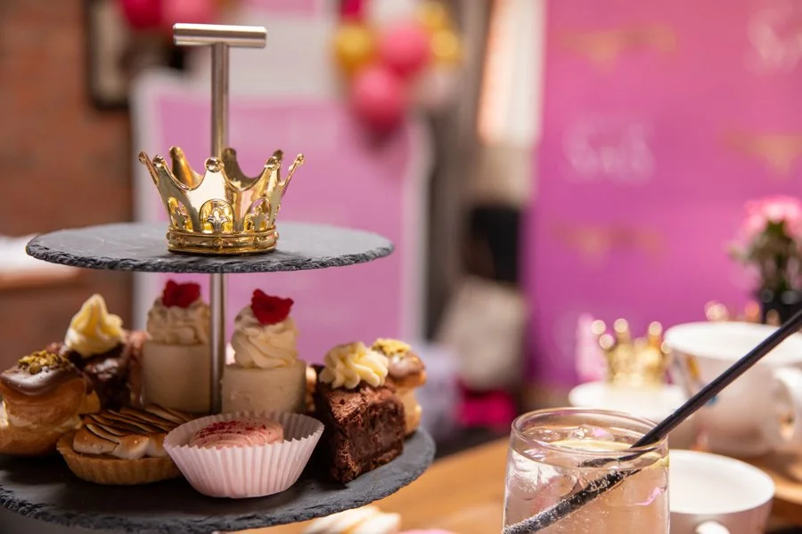 A two-tiered serving platter with assorted desserts, topped with a small gold crown, with a pink background featuring pink and yellow paper lanterns