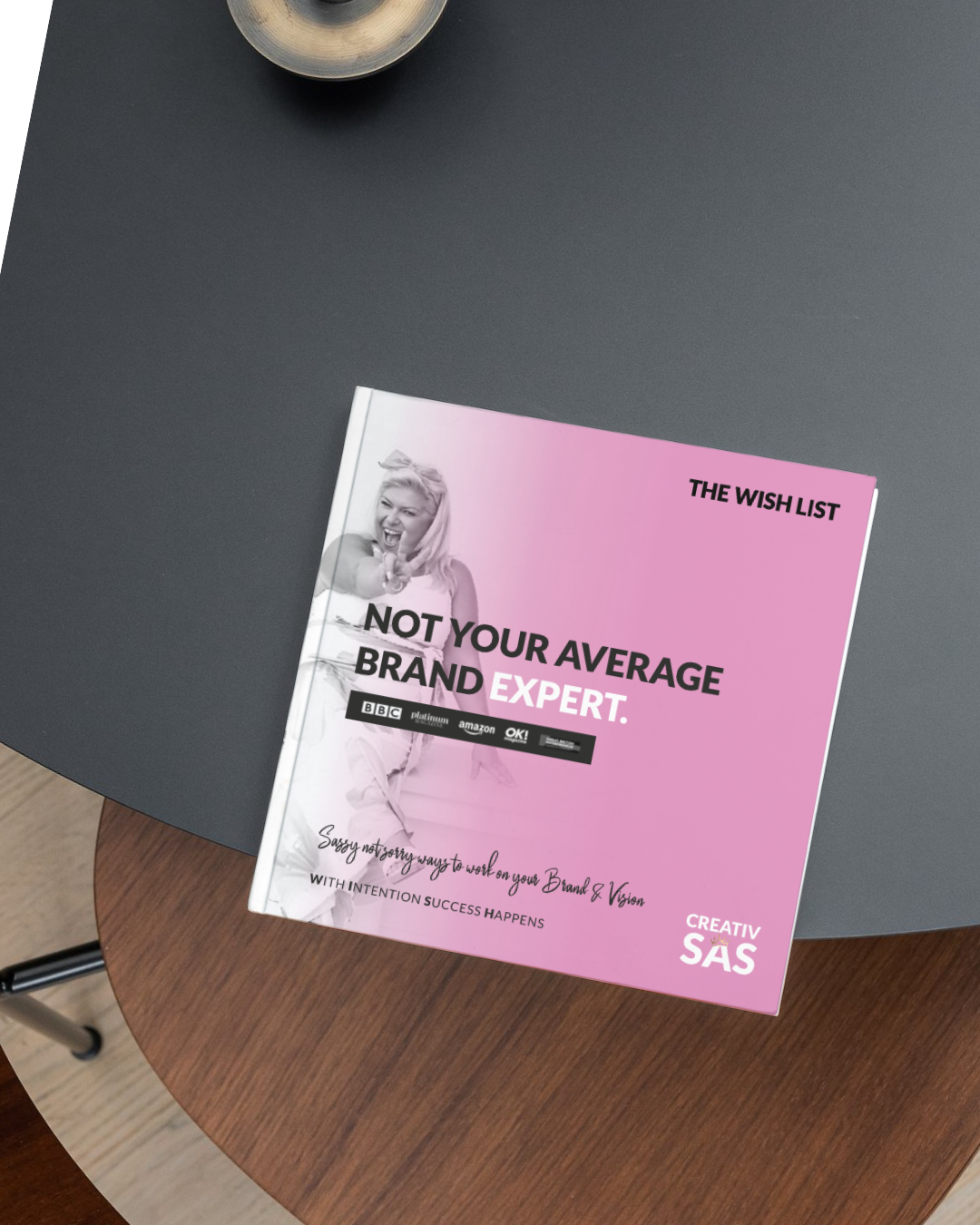 Pink and black booklet titled 'The Wish List' with the subtitle 'Not Your Average Brand Expert' and a black-and-white photo of a woman pointing and smiling, placed on a round wooden table with a dark gray surface nearby and a metallic cup at the top.