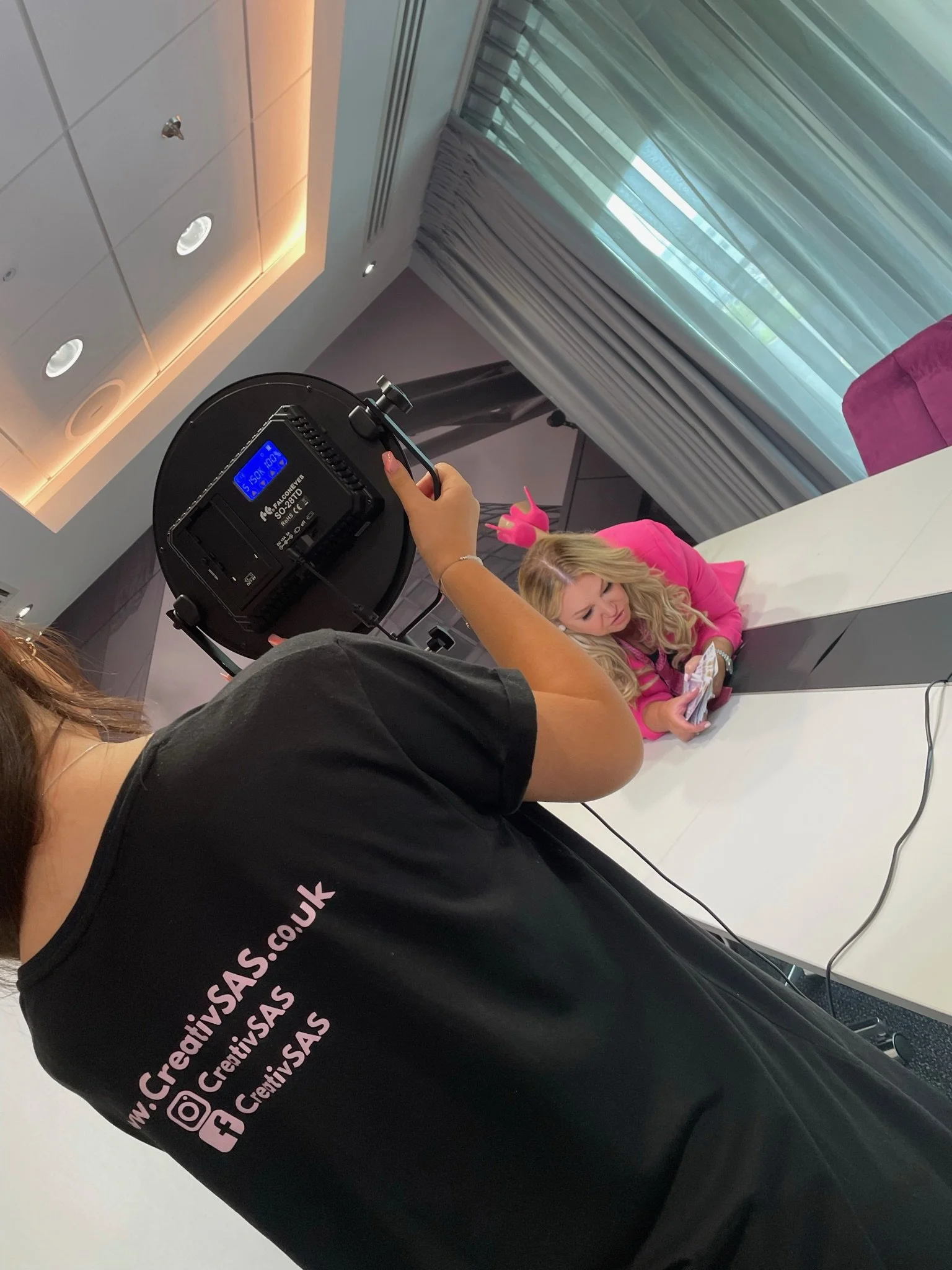 A woman holding a photography light is taking a photo of a young woman with blonde hair, wearing a pink jacket, lying on a white table, using a smartphone. The room has curtains, a window, and ceiling lights.