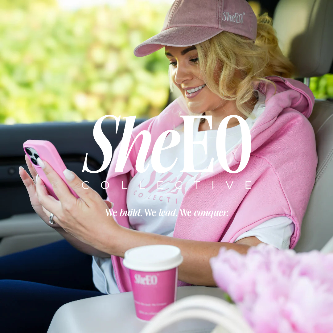 A woman wearing a pink cap and pink hoodie smiles while looking at her phone, seated in a vehicle with a pink coffee cup on her lap and blurred greenery outside.