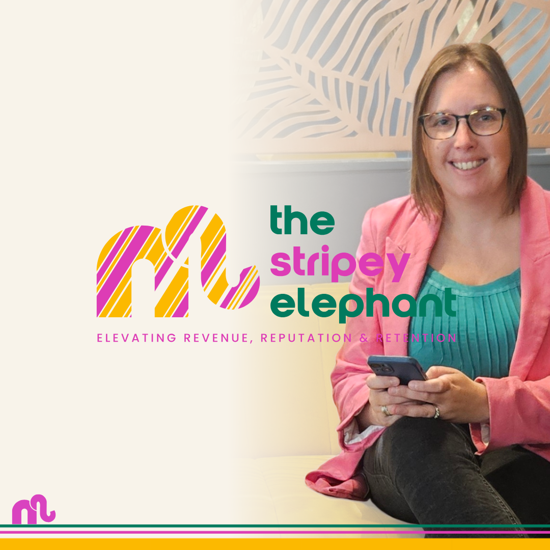 A woman with glasses in a pink blazer holding a smartphone, sitting on a yellow couch with a beige wall in the background decorated with a large abstract pattern.