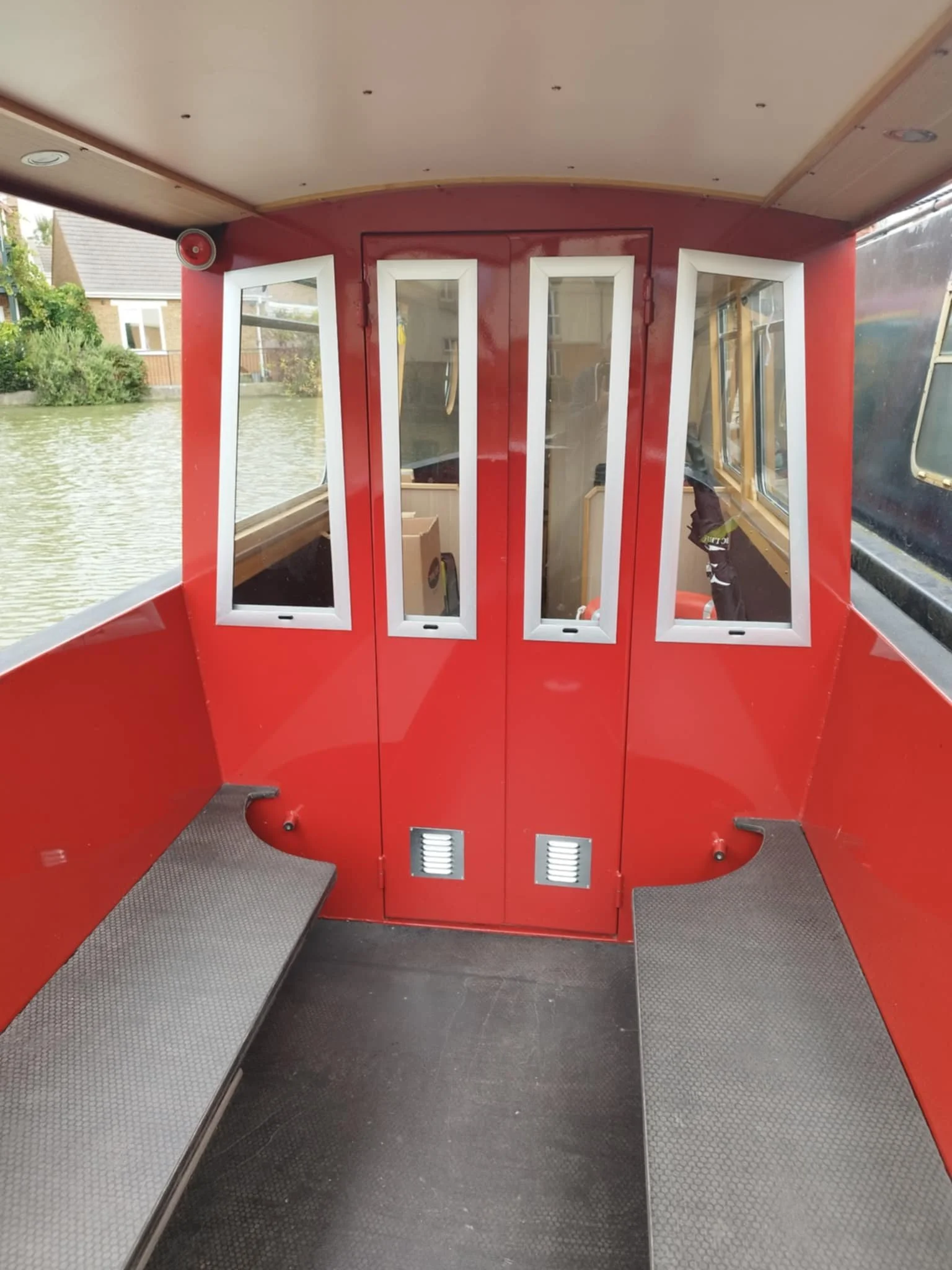 Bow of a red narrowboat boat with made to size tall windows, two silver benches, and water visible outside.