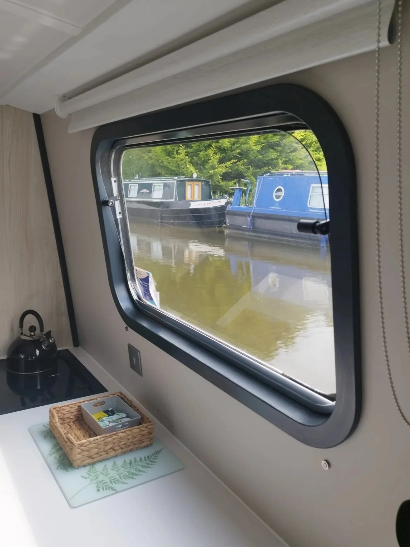 Inside view of a boat cabin window showing two moored narrowboats on the water with green trees in the background.