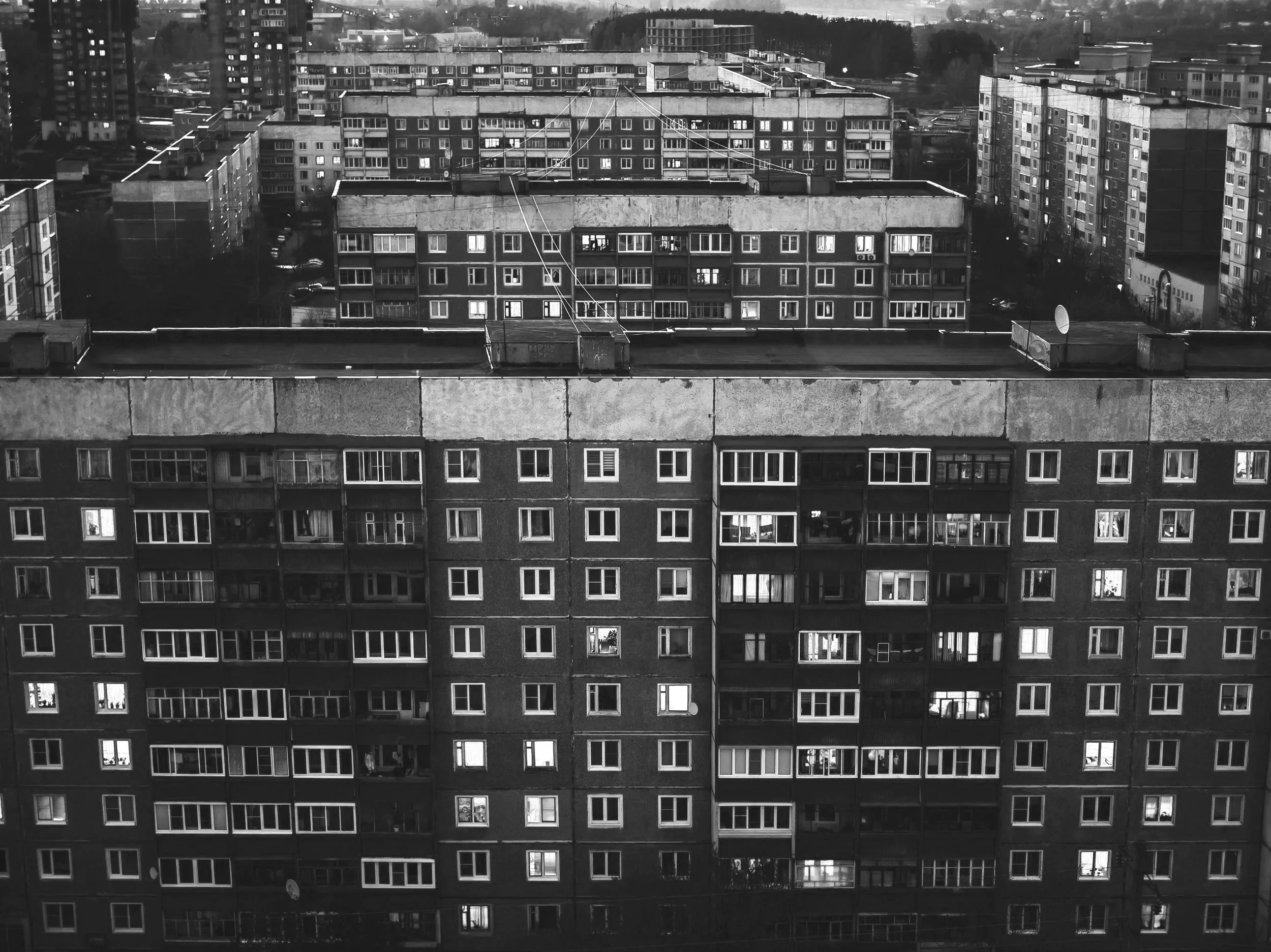 Black and white photograph of multiple apartment buildings in a city, with windows and balconies, with some windows lit up at dusk or night.
