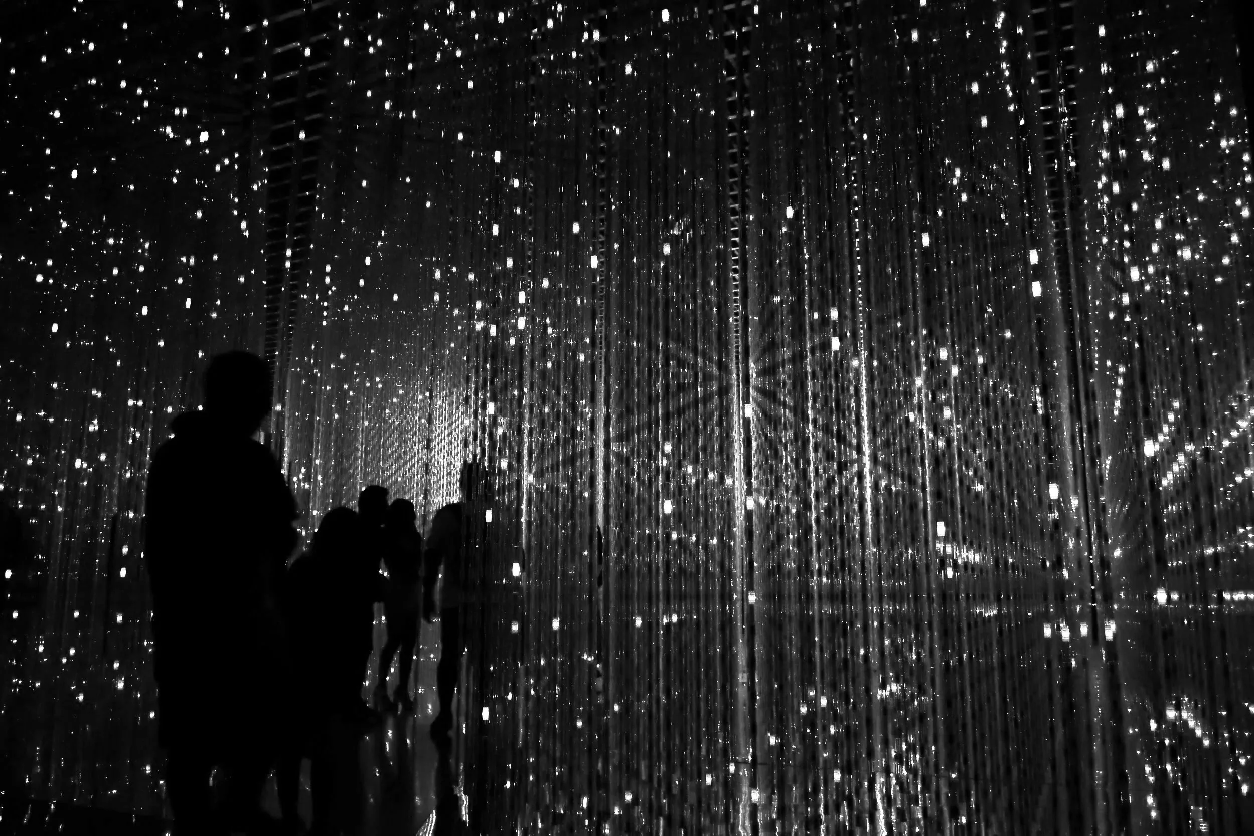 People viewing a dark room filled with hanging LED lights and mirrors creating a starry effect.