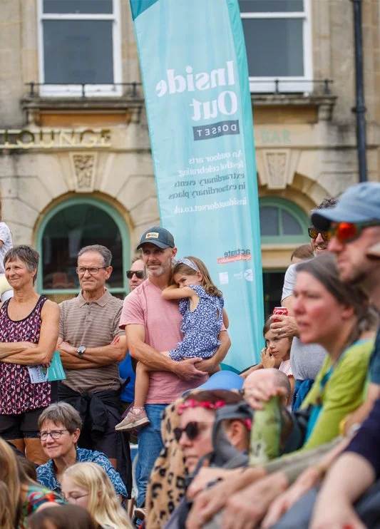 People gathered outdoors at an event, with a man holding a young girl, and a large turquoise banner reading 'Insidia' and 'Out' in the background.