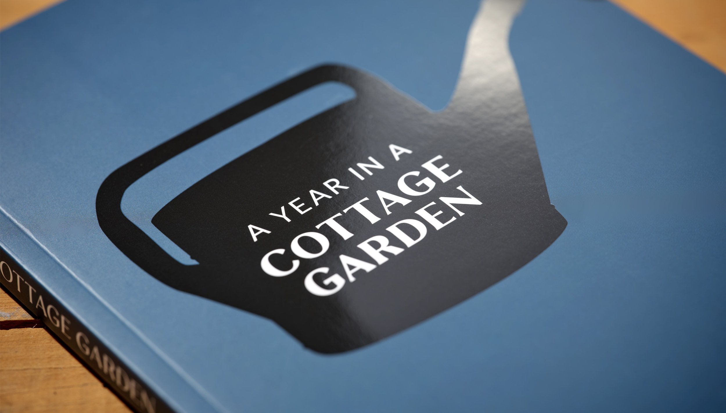 The cover of a book titled 'A Year in a Cottage Garden' with a large black silhouette of a watering can on a blue background.