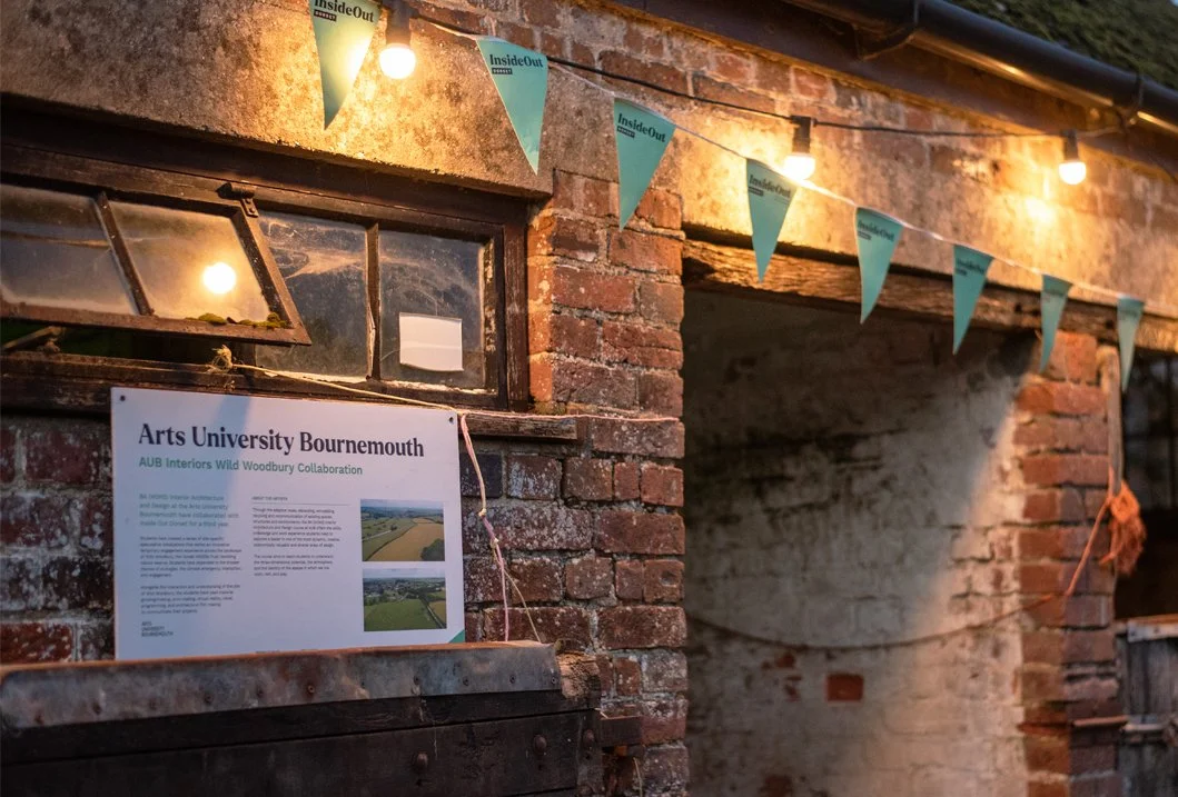 Outdoor brick wall decorated with string lights and turquoise pennant flags, a window with a sign about Arts University Bournemouth and woodbury collaboration, and a small open window with a slanted cover.