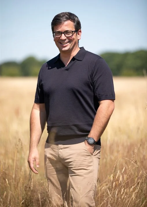 A man standing outdoors in a field of tall grass, smiling, wearing glasses, a black polo shirt, khaki pants, and a wristwatch.