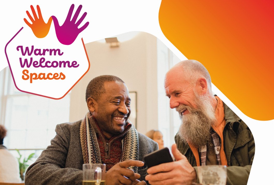 Two men smiling and looking at a smartphone at a social event with a banner reading 'Warm Welcome Spaces' with colorful hands above the text.