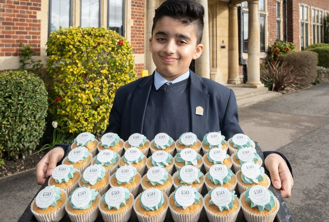 A young boy in a school uniform holding a tray of cupcakes decorated with a badge that reads "450". The background shows a brick building and greenery.