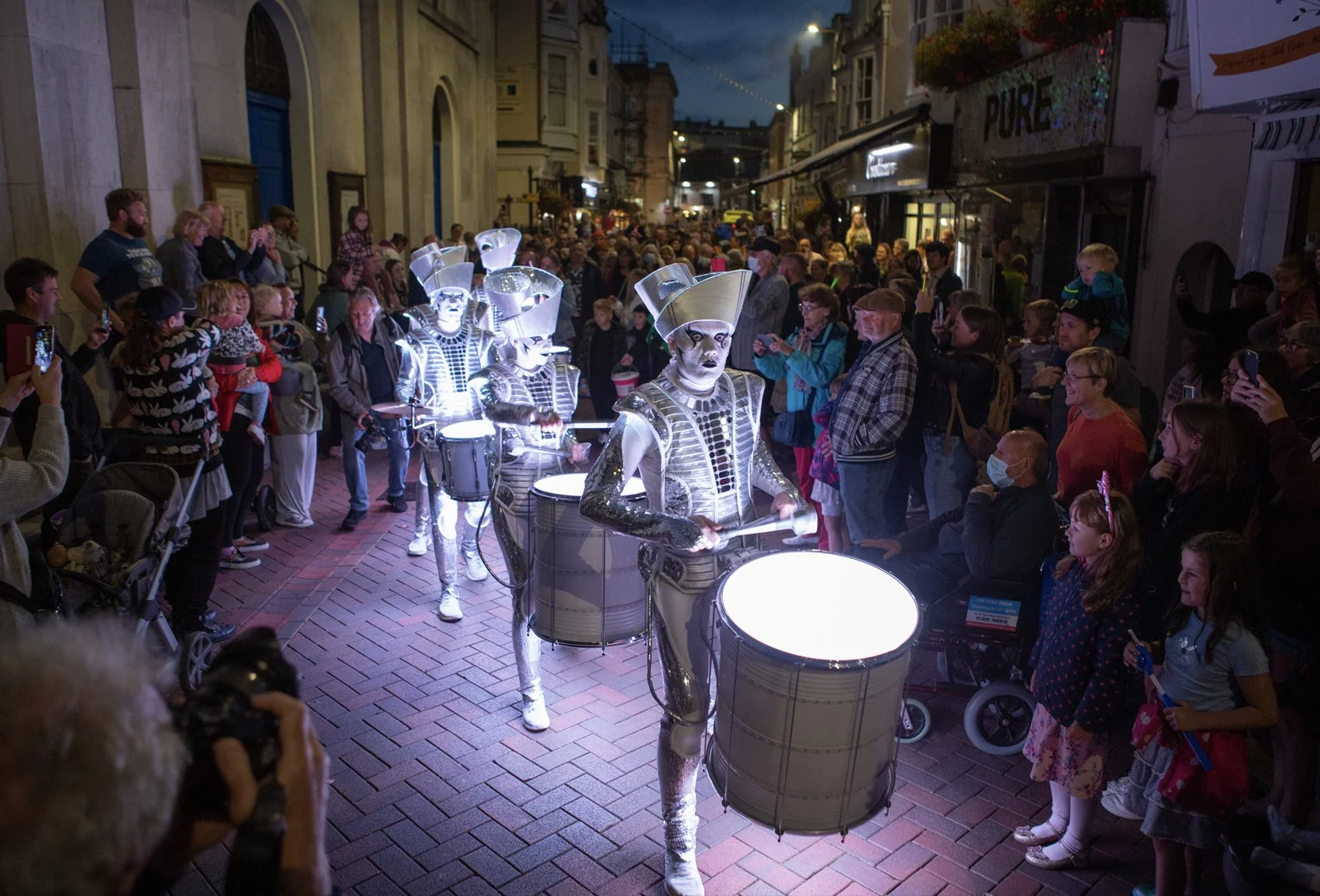 A street performer dressed as a futuristic drummer with shiny metallic costume and LED lights, performing for a crowd at night on a brick-paved street, surrounded by spectators including adults and children, many taking photos.