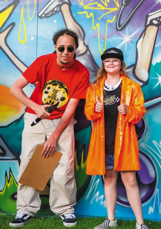 A young girl and a man posing in front of a colorful graffiti mural. The girl is wearing glasses, an orange shiny jacket, a black cap, and checkered shoes, giving a thumbs-up. The man is holding a microphone and a clipboard, wearing a red T-shirt with a yellow smiley face and the text "Save the World," beige pants, and sunglasses.