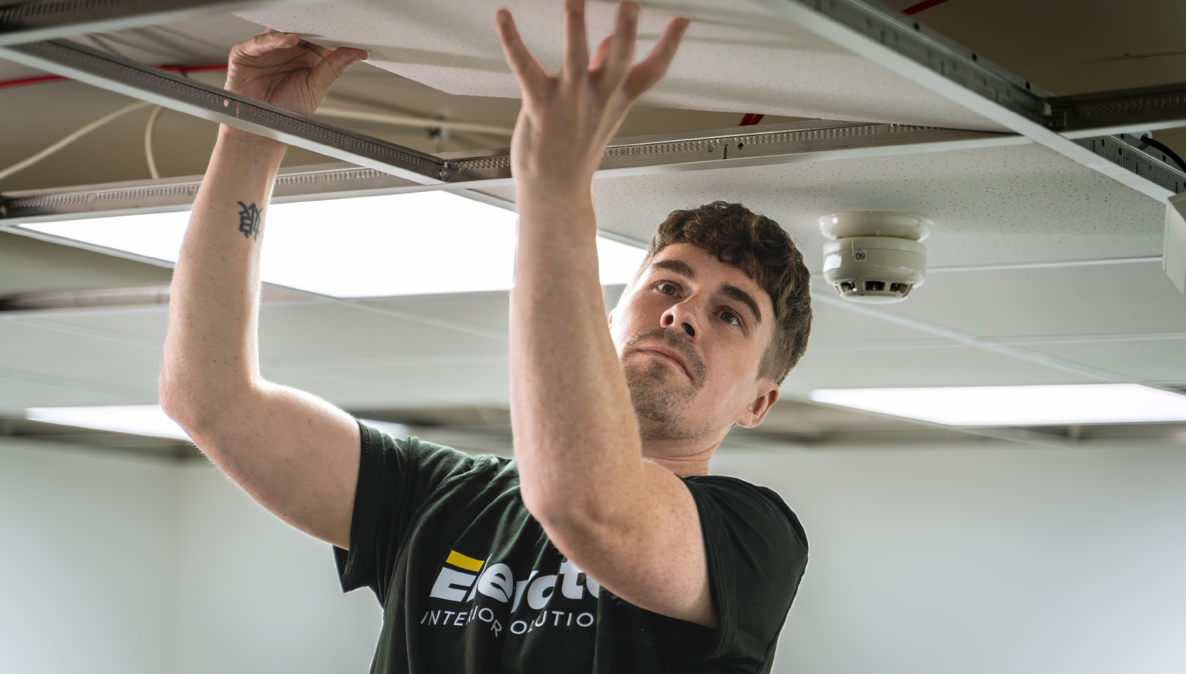 A man is installing or adjusting a ceiling-mounted device or panel, reaching up with both hands underneath a suspended ceiling.