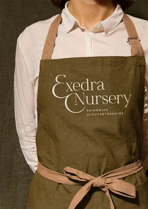 Person wearing a brown apron with 'Exedra Nursery, Painswick Gloucestershire' printed on it, over a white shirt.