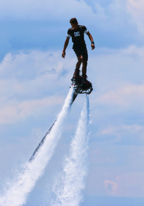 A person riding a water jetpack over the water with a blue sky and clouds in the background.