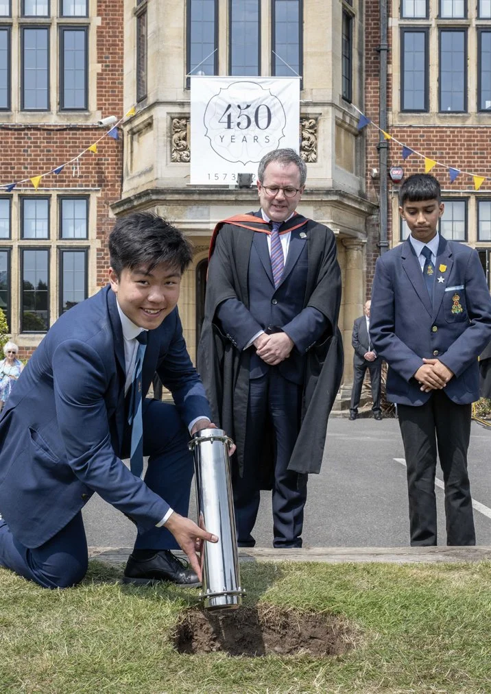 Two students in school uniforms and a person in academic regalia participate in a commemorative planting event outside a historic building with a banner celebrating 450 years, indicating a special anniversary celebration.