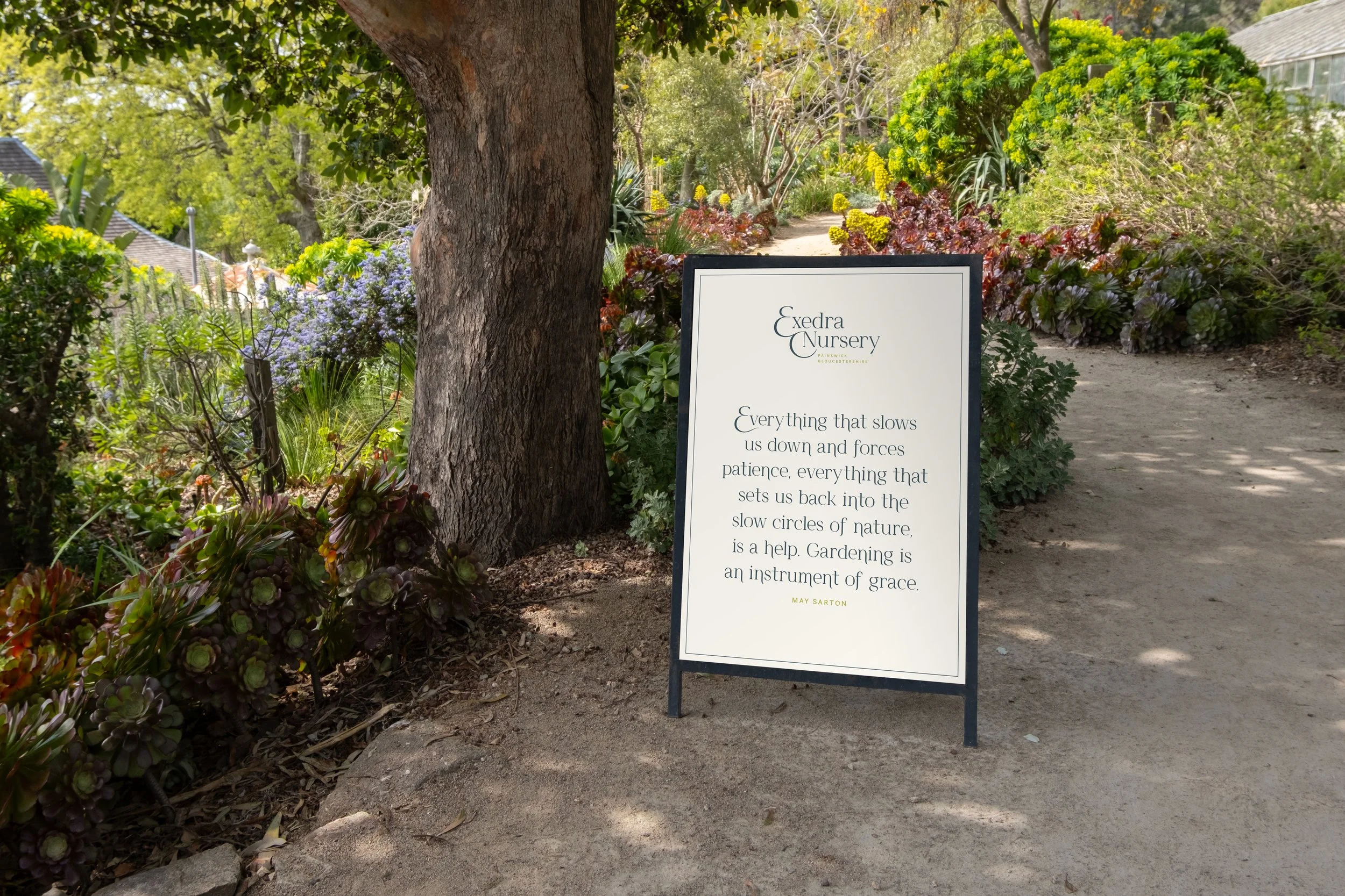 A signboard at the entrance of a garden pathway, with trees and colorful plants on either side.