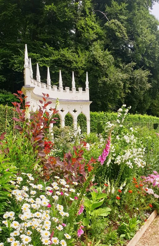 A garden with colourful flowers in the foreground and a small white castle-like structure with turrets in the background, surrounded by green trees.