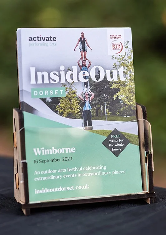 Brochure for InsideOut Dorset outdoor arts festival featuring a family performing an acro yoga pose outdoors, with promotional information about the event happening on September 16, 2023, in Wimborne, highlighting free family events.