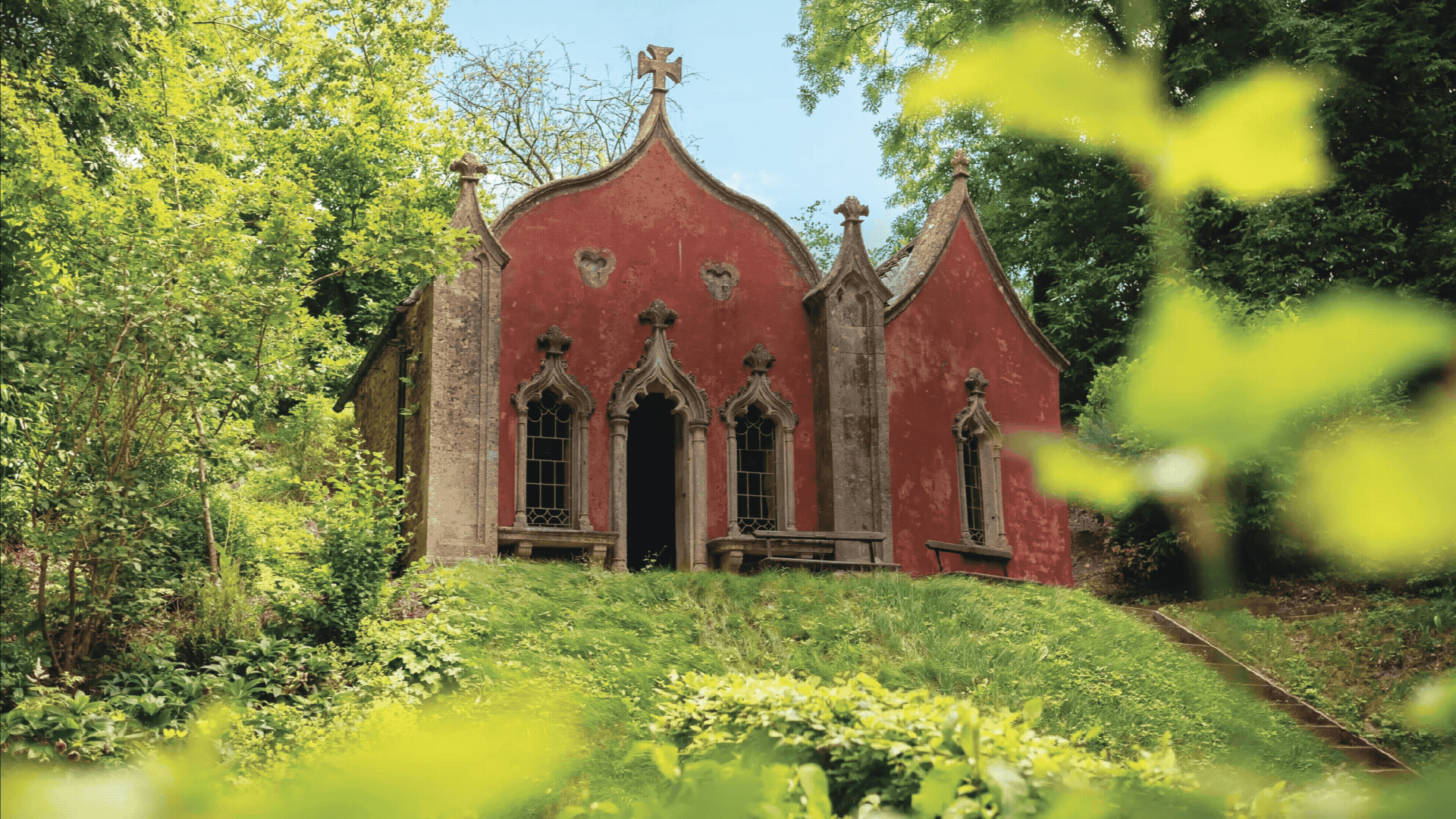 A red Gothic-style chapel surrounded by lush green trees and foliage, with a grassy slope leading up to it and a pathway on the right side With the logo of Rococo Graden.