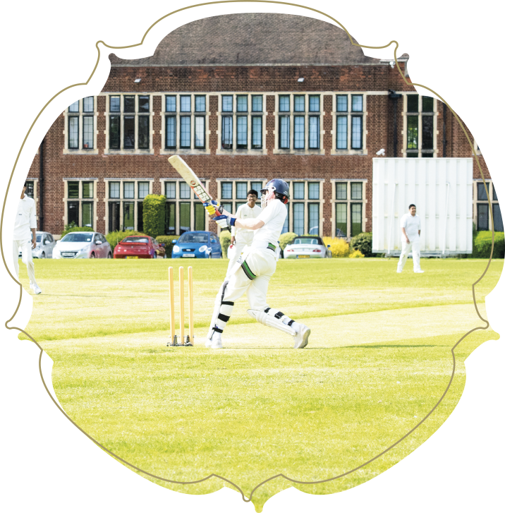 Cricket player batting on a grassy field with a large brick building in the background and other players around.