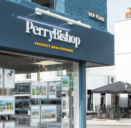 Perry Bishop real estate agency storefront with property listings displayed in the window and a street sign that reads Kew Place.