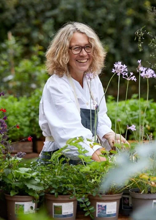A woman with curly blonde hair and glasses smiling while gardening among potted plants with purple flowers.