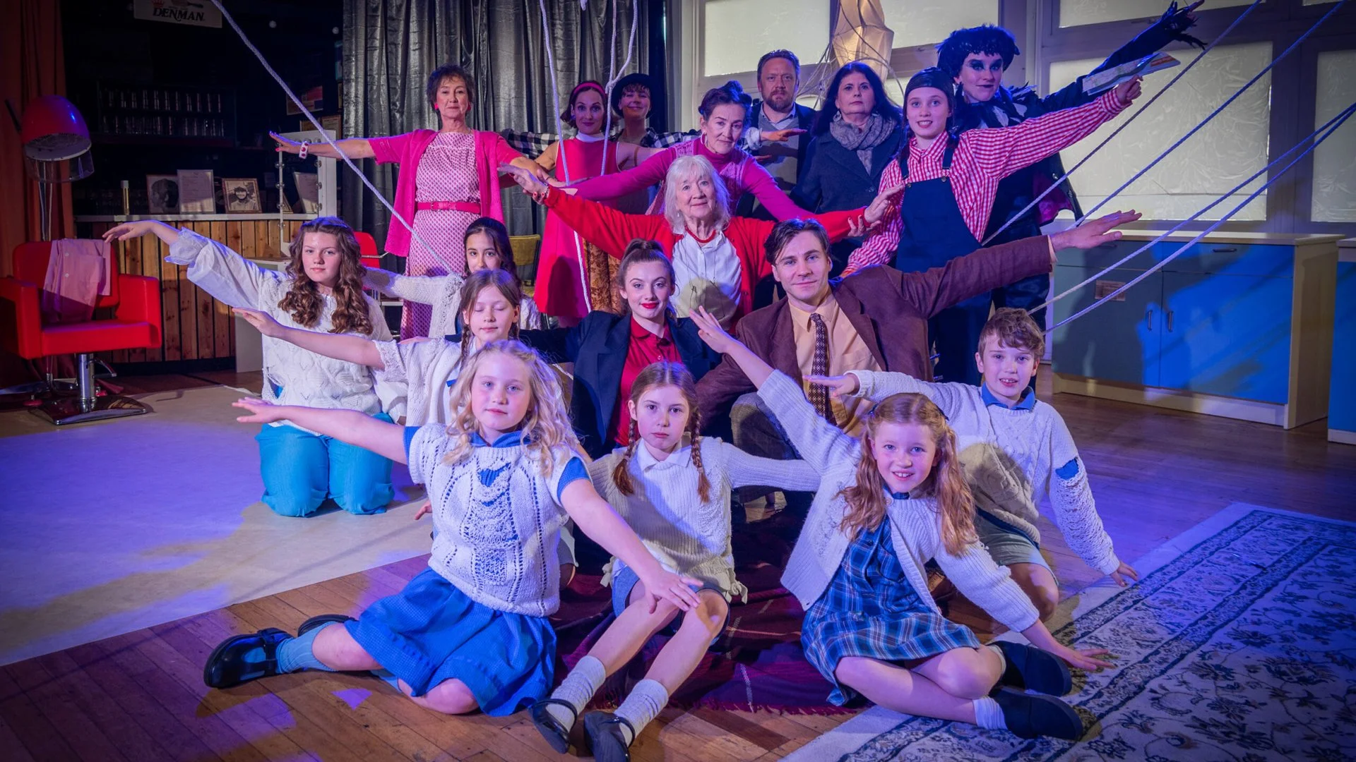 A group of children and adults dressed in vintage costumes posing on a stage with wooden floor, some acting as if performing a play or dance, with a backdrop of curtains and a window, decorated with props and furniture.