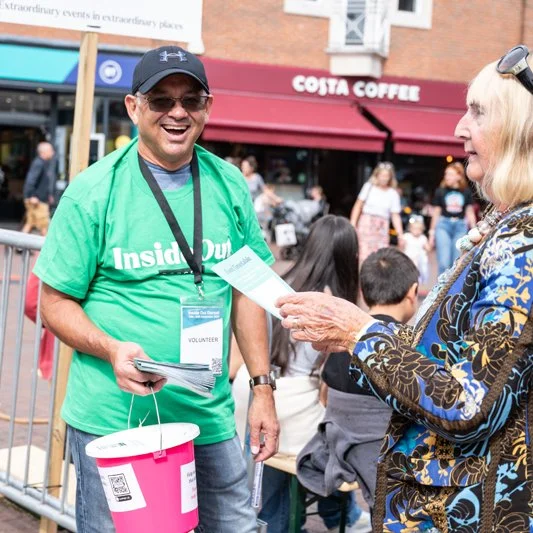 Volunteer man in green shirt smiling and talking to an older woman at an outdoor event, Costa Coffee sign visible in the background.