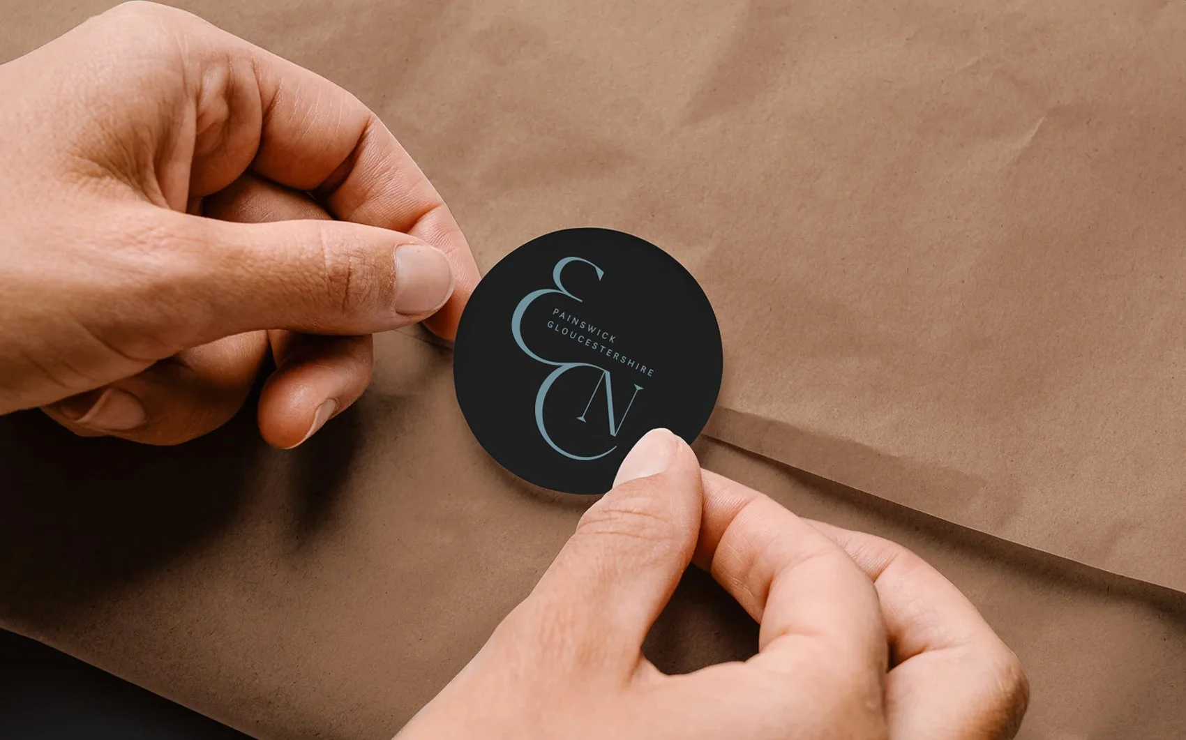 Person holding a round black sticker with blue text that says 'PAINSWICK GLOUCESTERSHIRE' and decorative initials 'C' and 'N'.