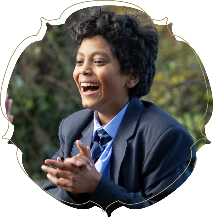 A young boy in a school uniform smiling and clapping outdoors with blurred trees in background.