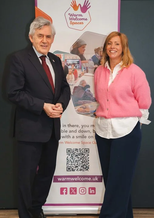A man in a dark suit and a woman in a pink cardigan posing together in front of a banner that reads 'Warm Welcome Spaces.' The banner features images of people and a QR code. The woman is smiling, and the man has his hands clasped in front of him.