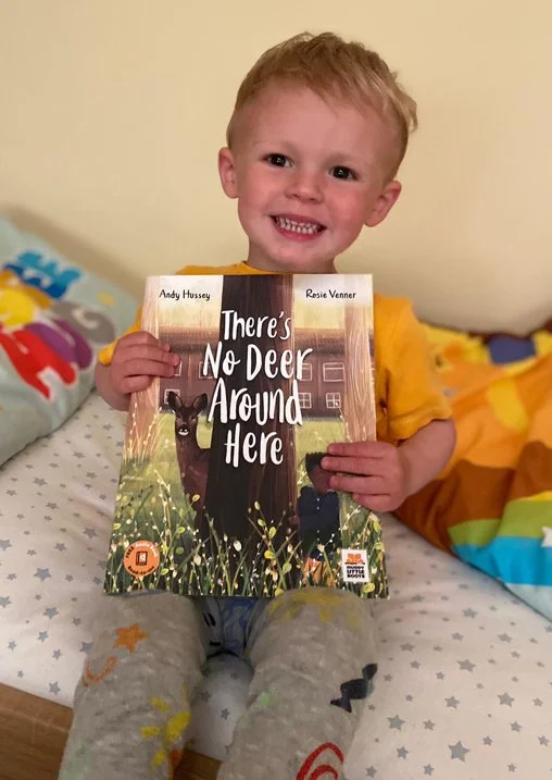 A young boy smiling and sitting on a bed, holding a children's book titled 'There's No Deer Around Here'.