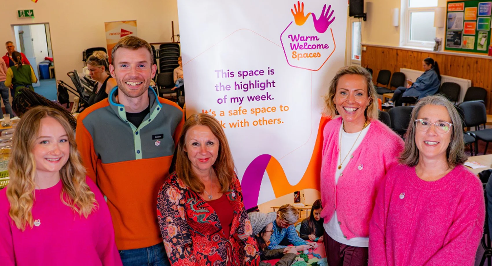 Group of five women and one man standing in front of a colorful sign reading 'Warm Welcome Spaces' with a friendly message about the space being safe for talking with others. The background shows a community center setting with additional people seated and engaging in activities.