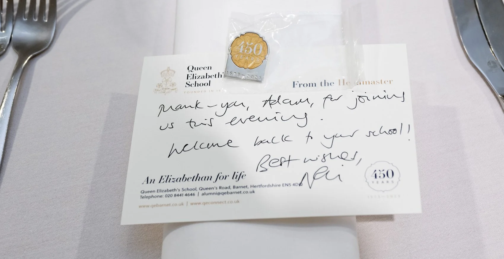 A note on a table from Queen Elizabeth's School with a gold 450 years anniversary coin in a plastic bag on top. The note thanks the recipient for joining that evening and welcoming them back to school, signed with best wishes.